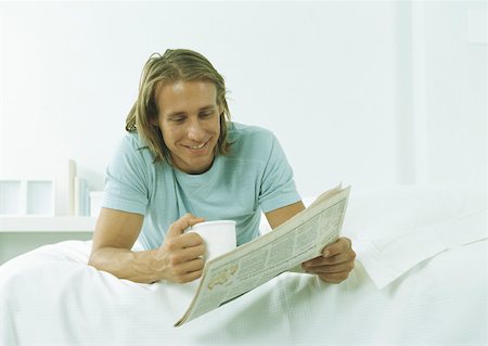 Young man lying on stomach with mug in one hand and newspaper in other Stock Photo - Premium Royalty-Free, Code: 632-01151486