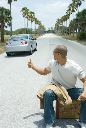 Male hitchhiker sitting on suitcase on road, looking over shoulder at passing car Foto de stock - Sin royalties Premium, Código: 632-01785257