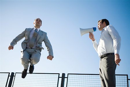 Businessman shouting into megaphone, colleague jumping in the air, low angle view Stock Photo - Premium Royalty-Free, Code: 632-01828133