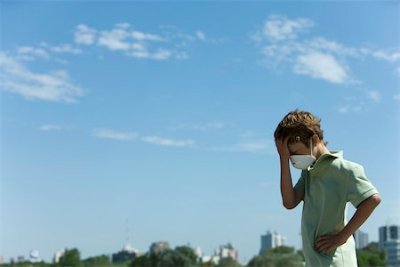 Boy standing outdoors wearing pollution mask, holding head, city in background Stock Photo - Premium Royalty-Free, Code: 632-02745095