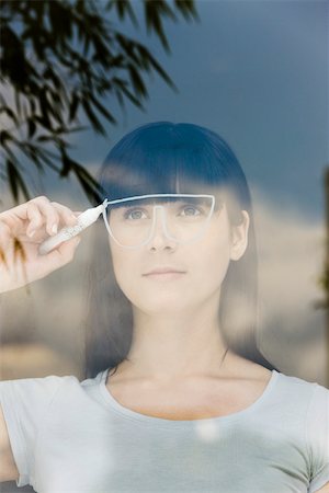 Young woman drawing a pair of glasses on a window pane, looking through window Foto de stock - Royalty Free Premium, Número: 632-02745320