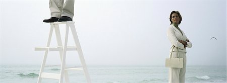 Woman on beach with folded arms watching man standing on top of ladder, cropped Stock Photo - Premium Royalty-Free, Code: 632-03083081