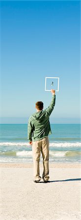 Man on beach holding up picture frame capturing image of gull flying against blue sky Stock Photo - Premium Royalty-Free, Code: 632-03500760