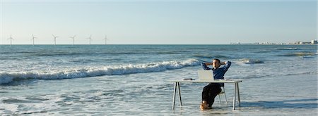 simsearch:632-03500786,k - Man relaxing at desk set at water's edge on beach, wind turbines on horizon Stock Photo - Premium Royalty-Free, Code: 632-03500777