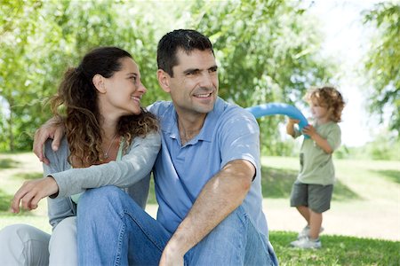 simsearch:695-03379156,k - Couple relaxing together outdoors, woman looking over shoulder at son playing in background Stock Photo - Premium Royalty-Free, Code: 632-03516982