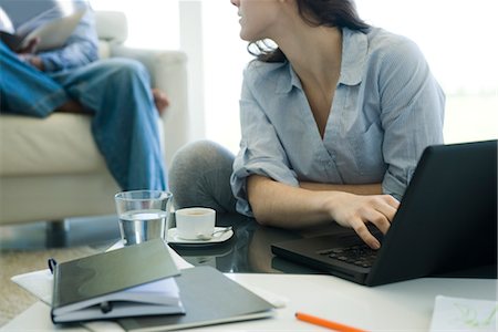 Femme utilisant un ordinateur portable dans le salon, regardant par-dessus son épaule à l'homme assis en arrière-plan Photographie de stock - Premium Libres de Droits, Code: 632-03779712