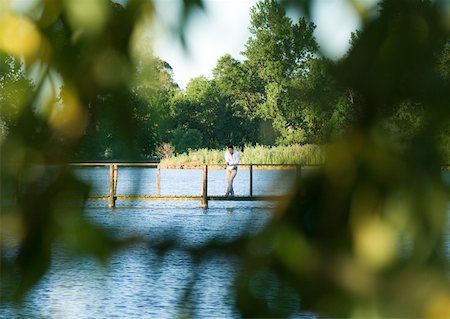 simsearch:632-05554100,k - Man standing on footbridge over river, in distance, seen through leaves in blurred foreground Foto de stock - Royalty Free Premium, Número: 633-01273471