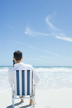 simsearch:633-01713848,k - Homme assis dans la chaise, pliante brandissant un téléphone cellulaire, sur la plage Photographie de stock - Premium Libres de Droits, Code: 633-01574610