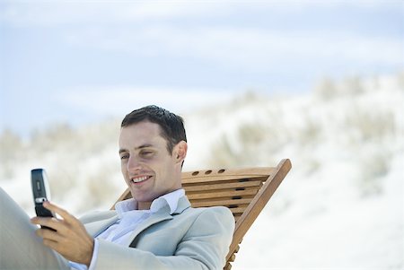 simsearch:633-01574593,k - Businessman sitting in lounge chair on beach, looking down at cell phone, smiling Stock Photo - Premium Royalty-Free, Code: 633-01574626