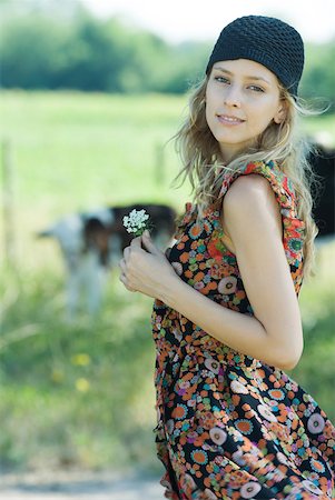 simsearch:633-01715239,k - Jeune femme debout avec une fleur à la main dans un cadre champêtre, souriant à la caméra Photographie de stock - Premium Libres de Droits, Code: 633-01714154