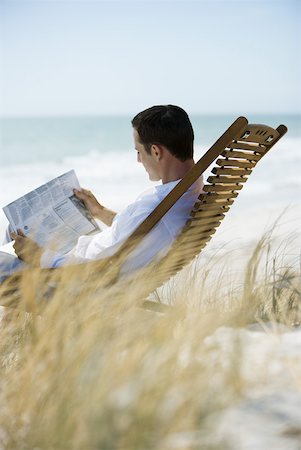 Young man sitting in deck chair on beach, reading newspaper Stock Photo - Premium Royalty-Free, Code: 633-01714425