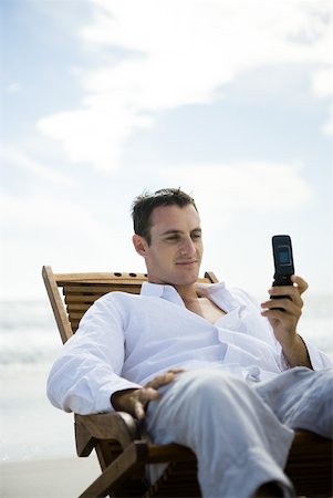 Young man sitting in chair on beach, using cell phone Stock Photo - Premium Royalty-Free, Code: 633-01714442