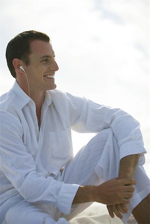 Young man sitting with knee up, listening to earphones, smiling, looking away Stock Photo - Premium Royalty-Free, Code: 633-01714465