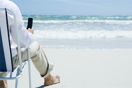simsearch:632-03500777,k - Man sitting in folding chair on beach, holding cell phone, cropped, rear view Foto de stock - Royalty Free Premium, Número: 633-01713851