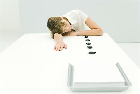 simsearch:633-02417737,k - Woman resting head on table, lined up stones leading to stack of paper in tray Fotografie stock - Premium Royalty-Free, Codice: 633-02231713