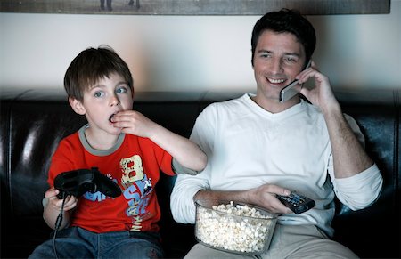 Father talking on cell phone while boy is playing a video game Stock Photo - Premium Royalty-Free, Code: 644-01436473