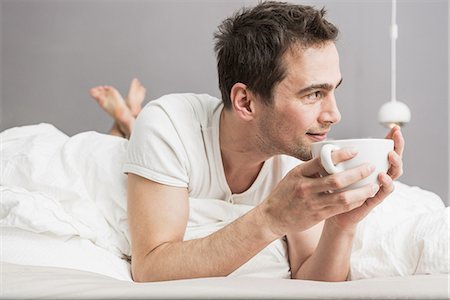 pyjamas (sleepwear with pants) - Mid adult man lying on front on bed, holding coffee cup Stock Photo - Premium Royalty-Free, Code: 649-06844756