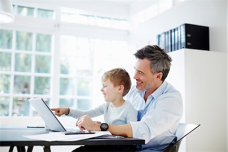 Man sitting at desk with son on his knee with laptop computer and paperwork Stock Photo - Premium Royalty-Free, Code: 649-07064544