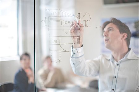 Man drawing architectural plans on glass wall, colleagues in background Stock Photo - Premium Royalty-Free, Code: 649-07238400