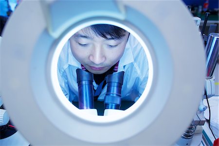 Worker at small parts manufacturing factory in China looking through microscope Stock Photo - Premium Royalty-Free, Code: 649-07436359