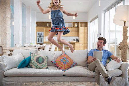 Girl jumping mid air from living room sofa whilst father uses digital tablet Photographie de stock - Premium Libres de Droits, Code: 649-08060388