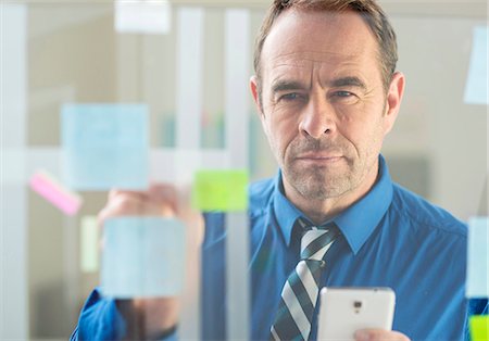 Businessman with smartphone sticking notes on office glass wall Stock Photo - Premium Royalty-Free, Code: 649-08084886
