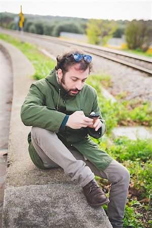devices - Mid adult man sitting on wall next to railway track using smartphone Foto de stock - Sin royalties Premium, Código: 649-08125700