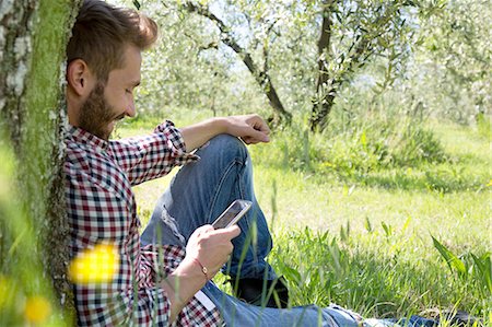 Young man sitting leaning against tree using smartphone looking down smiling Stock Photo - Premium Royalty-Free, Code: 649-08239066