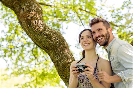 Young couple underneath tree holding camera looking away smiling Stock Photo - Premium Royalty-Free, Code: 649-08239092