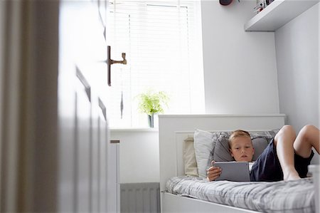 View through doorway of boy lying on bed looking down at digital tablet Stock Photo - Premium Royalty-Free, Code: 649-08306390