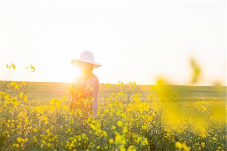 simsearch:649-09176954,k - Mid adult woman in canola field wearing sunhat smiling, focus on background, lens flare Foto de stock - Sin royalties Premium, Código: 649-08306675