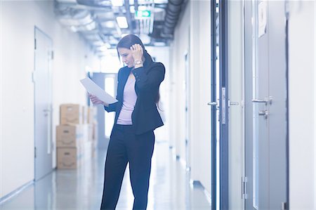 simsearch:400-08343539,k - Young woman standing in office corridor looking down at paperwork scratching head Photographie de stock - Premium Libres de Droits, Code: 649-08329102