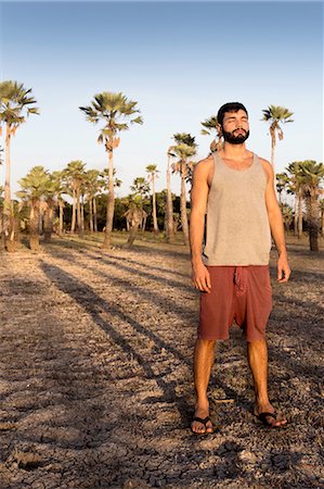 Full length front view of young man standing in front of palm trees casting shadow looking away, Taiba, Ceara, Brazil Stock Photo - Premium Royalty-Free, Code: 649-08381497