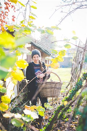 simsearch:649-08563716,k - View through leaves of young woman in garden wearing knit hat holding wickerwork basket looking at camera smiling Foto de stock - Sin royalties Premium, Código: 649-08422693