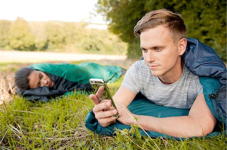 simsearch:649-08422994,k - Young man lying sleeping bag texting on smartphone in field Photographie de stock - Premium Libres de Droits, Code: 649-08423003
