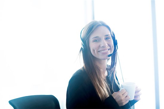 Young woman wearing telephone headset holding coffee cup looking at camera smiling Stock Photo - Premium Royalty-Free, Image code: 649-08423017