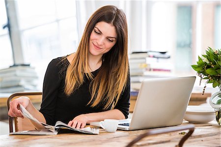 Young woman in city apartment reading magazine whilst working on laptop Stock Photo - Premium Royalty-Free, Code: 649-08544171