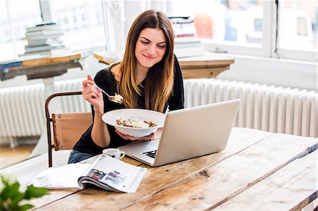 Young woman in city apartment eating muesli breakfast whilst reading  laptop Stock Photo - Premium Royalty-Free, Code: 649-08544172
