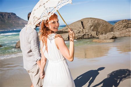 simsearch:649-08969763,k - Couple holding hands, walking on coastline holding umbrella looking over shoulder at camera smiling Fotografie stock - Premium Royalty-Free, Codice: 649-08543883