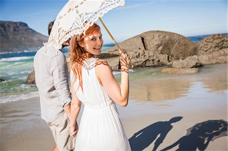 simsearch:6113-07992154,k - Couple holding hands, walking on coastline holding umbrella looking over shoulder at camera smiling Foto de stock - Sin royalties Premium, Código: 649-08543883