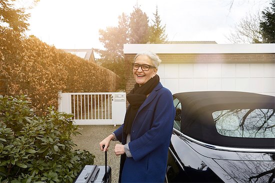 Woman near car on driveway holding suitcase looking at camera smiling Stock Photo - Premium Royalty-Free, Image code: 649-08576770