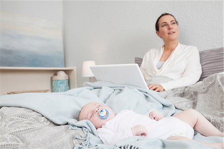 simsearch:649-08661252,k - Mother sitting in bed using laptop while baby boy sleeps beside her Foto de stock - Royalty Free Premium, Número: 649-08661257