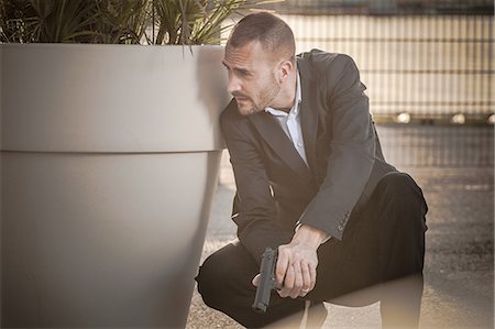 Man in business attire behind plant pot holding handgun Stock Photo - Premium Royalty-Free, Code: 649-08714602