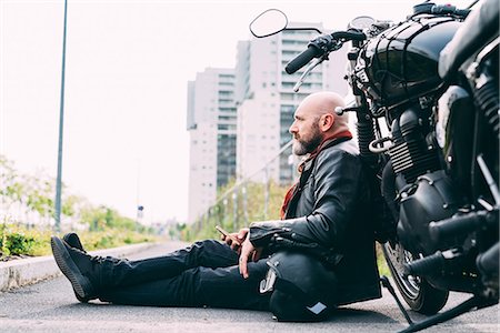 Mature male motorcyclist sitting on roadside leaning on motorcycle with smartphone Stock Photo - Premium Royalty-Free, Code: 649-08714733