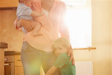 simsearch:614-08641537,k - Boy hugging mother's leg while she carries his baby brother in kitchen Photographie de stock - Premium Libres de Droits, Code: 649-08901453