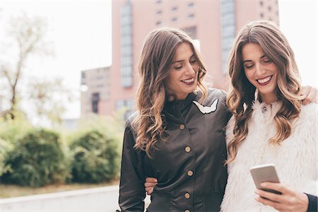 simsearch:649-08923050,k - Twin sisters, walking outdoors, looking at smartphone, smiling Stock Photo - Premium Royalty-Free, Code: 649-08923058