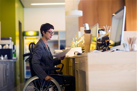 eyeglasses for female workers - Woman in wheelchair, sitting at desk, looking at document Photographie de stock - Premium Libres de Droits, Code: 649-08923718