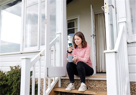 simsearch:649-08859802,k - Young woman sitting on porch stairs with coffee cup looking at smartphone Stock Photo - Premium Royalty-Free, Code: 649-08951073