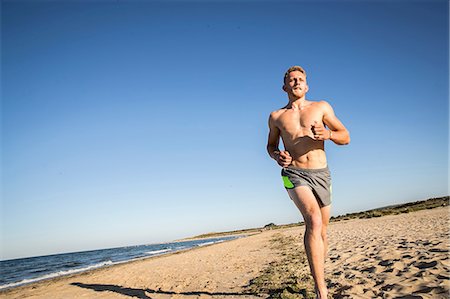 simsearch:649-08238188,k - Bare chested young male runner running along beach against blue sky Foto de stock - Sin royalties Premium, Código: 649-08988271