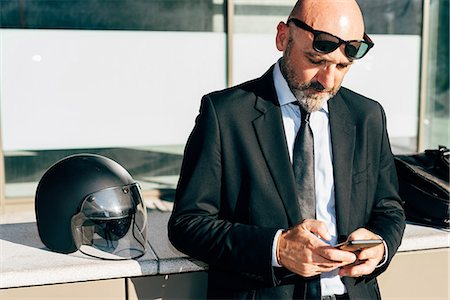 Mature businessman standing outdoors, using smartphone, motorcycle helmet on wall beside him Stock Photo - Premium Royalty-Free, Code: 649-09061328